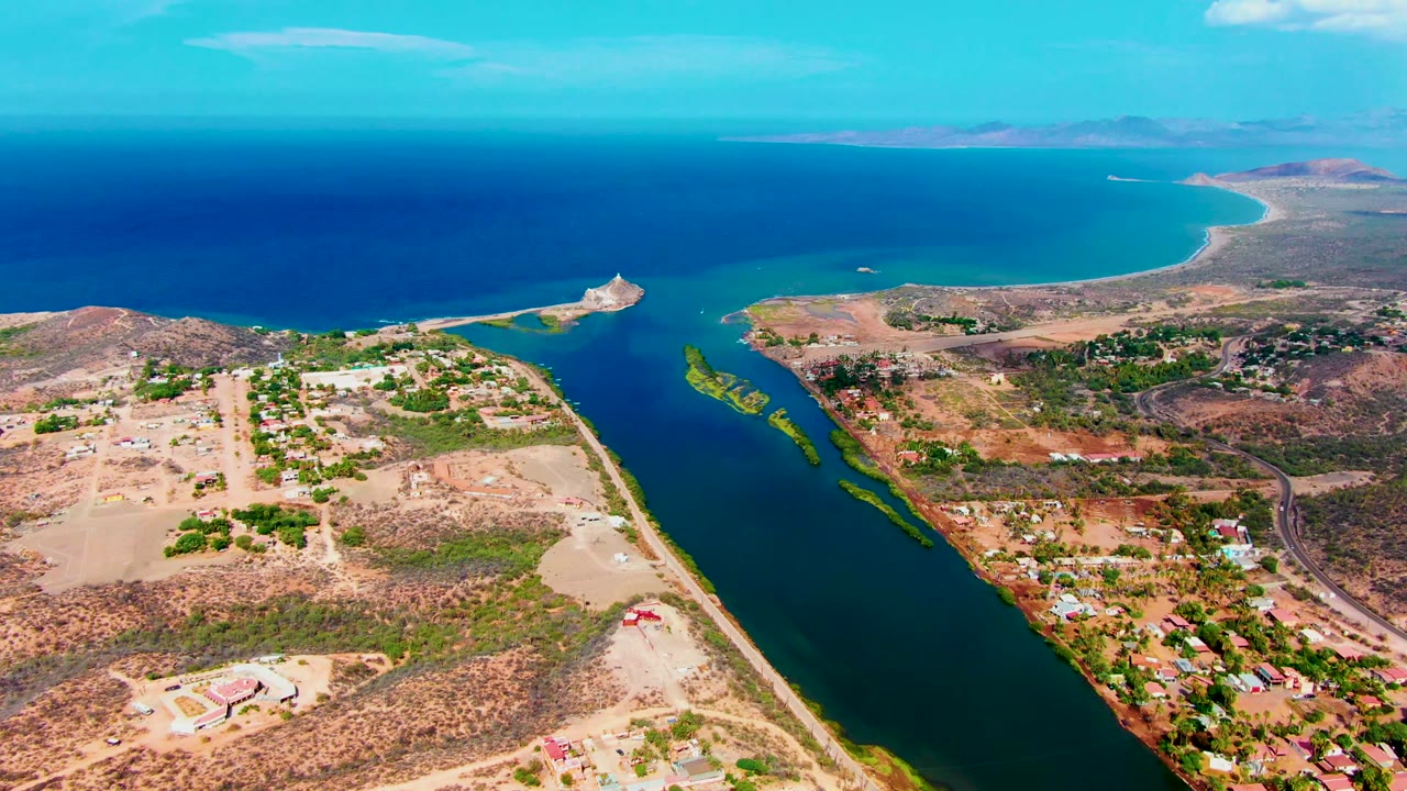 Drone-style aerial view over Mulege, Baja California Sur, showing the canal, pale dirt roads, clustered greenery, small buildings, ocean beyond, and no visible people.