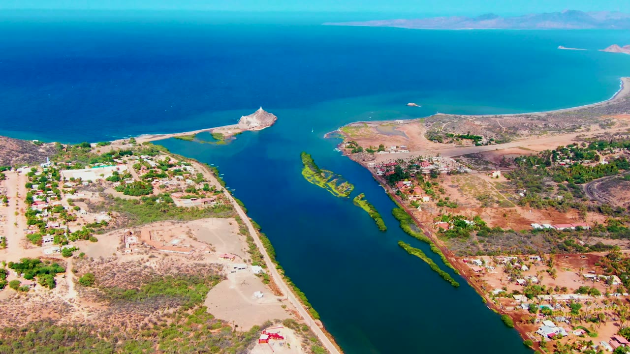 Opening aerial frame over Mulege, Baja California Sur, showing a narrow blue inlet, arid shoreline, scattered buildings, shore roads, distant islands, and no visible people.
