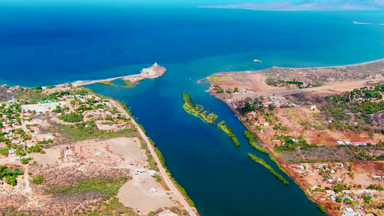 Drone-height view of turquoise coastal water and dry shorelines with roads and buildings.