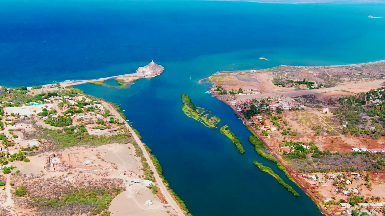 Mulege aerial frame looking across the blue inlet toward a small rocky point, with shoreline roads, dry lots, vegetation strips, scattered buildings, and no visible people.