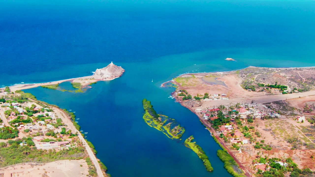 Oblique aerial view of Mulege, Baja California Sur, with turquoise water near the sea, a darker blue channel, narrow green islands, roads, low buildings, dry lots, and no visible people.