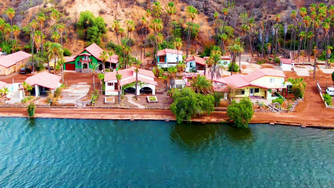 Aerial frame from Mulege, Baja California Sur showing blue-green water in front of small shoreline homes, palm trees, dry hills, and no visible people.