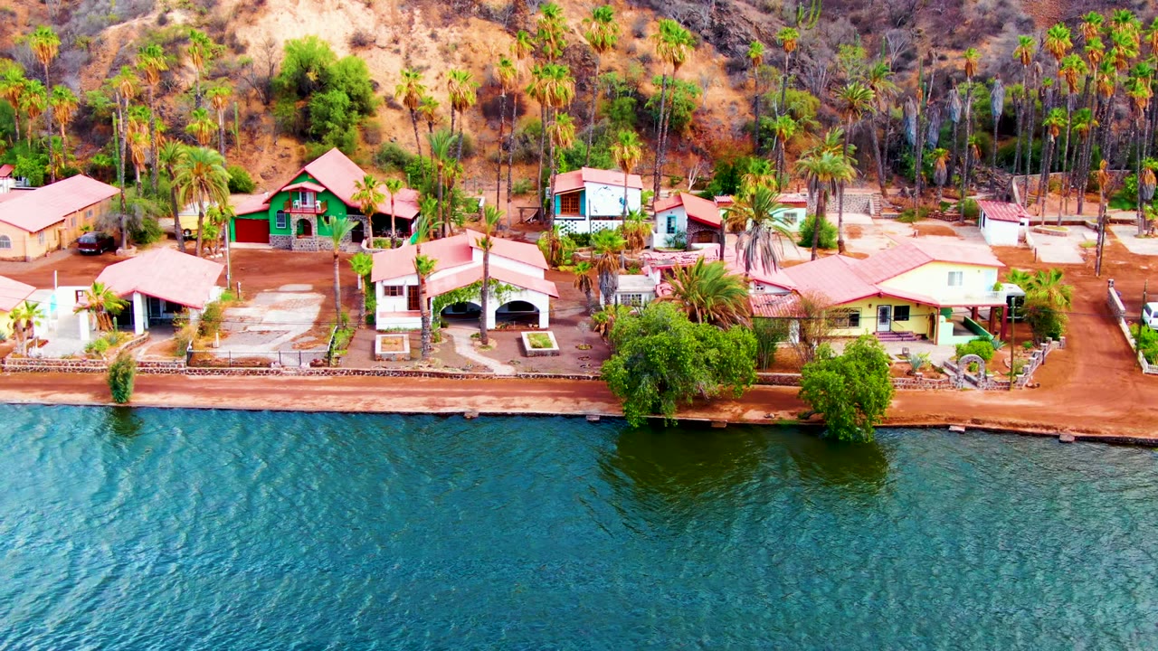 Aerial view in Mulege, Baja California Sur with waterfront homes, tall palms, a narrow shore edge, textured water, hillside brush, and no visible people.
