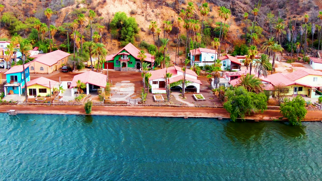 Aerial frame over Mulege, Baja California Sur showing blue-green rippling water, a seawall, colorful houses with red tile roofs, palm trees, dry hills, and no visible people.