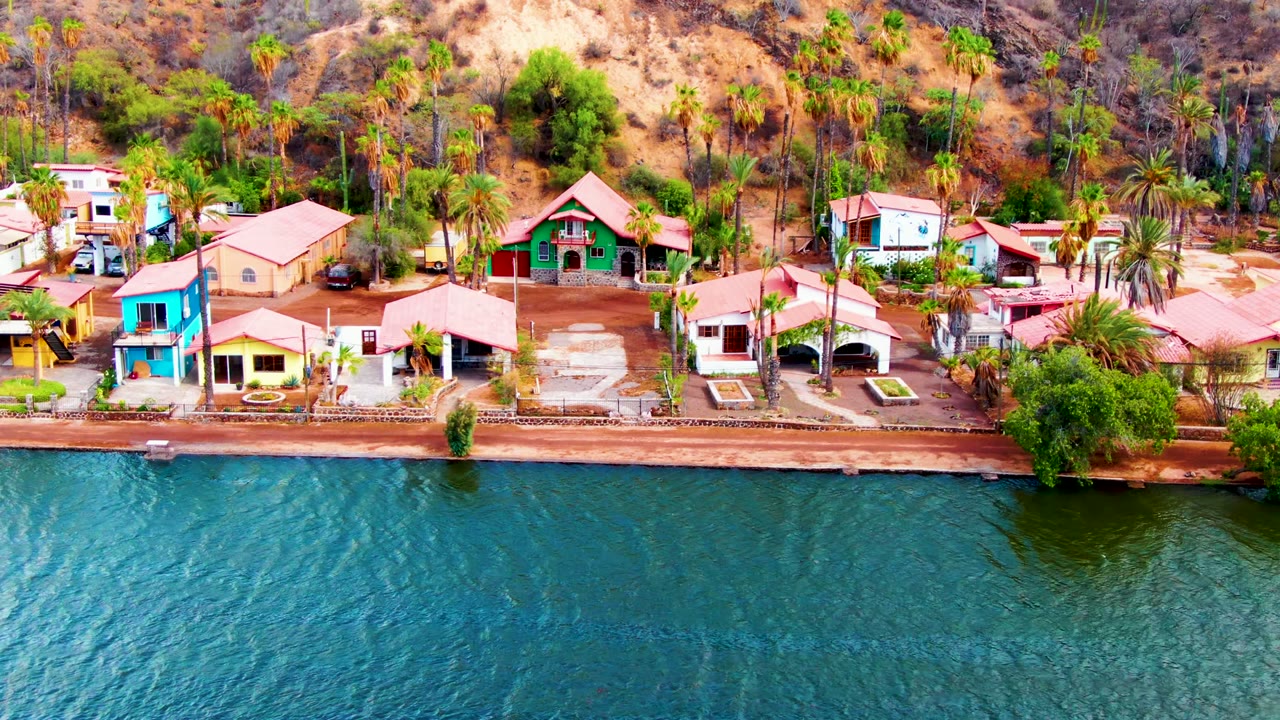 Aerial frame over Mulege, Baja California Sur showing the house row centered more tightly, with blue water below, palms between buildings, tan hillside behind, and no visible people.
