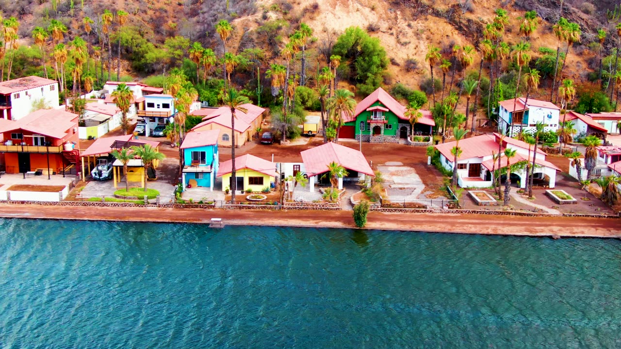 Aerial frame in Mulege, Baja California Sur showing blue water in front of red-roofed waterfront houses, palms, and a dry hillside, with no visible people.