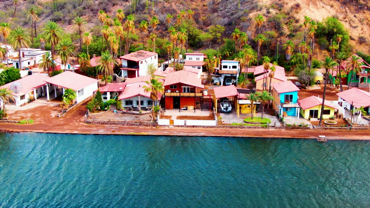 Aerial frame over blue-green water toward Mulege shoreline homes with red roofs, palm trees, dry hillside brush, and no visible people.