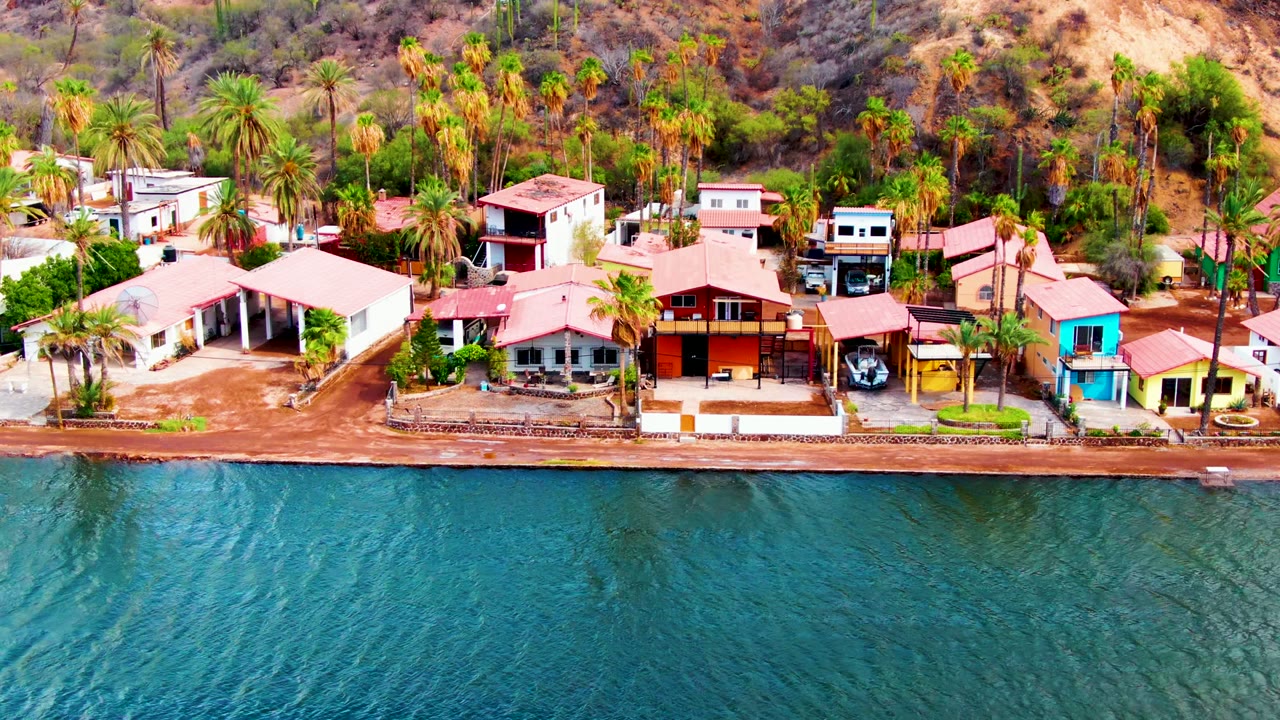 Aerial frame of Mulege waterfront homes and palms, with blue-green water below, red and white roofs along the shore, dry hillside behind, and no visible people.