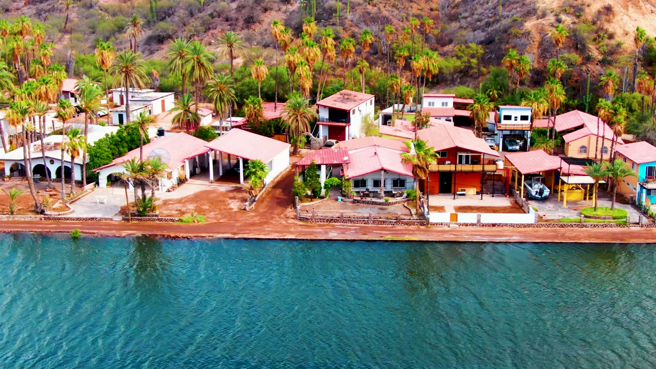 Aerial frame from Mulege, Baja California Sur showing red-roofed waterfront buildings, palms, sandy shoreline, dry hillside, and rippling blue-green water with no visible people.