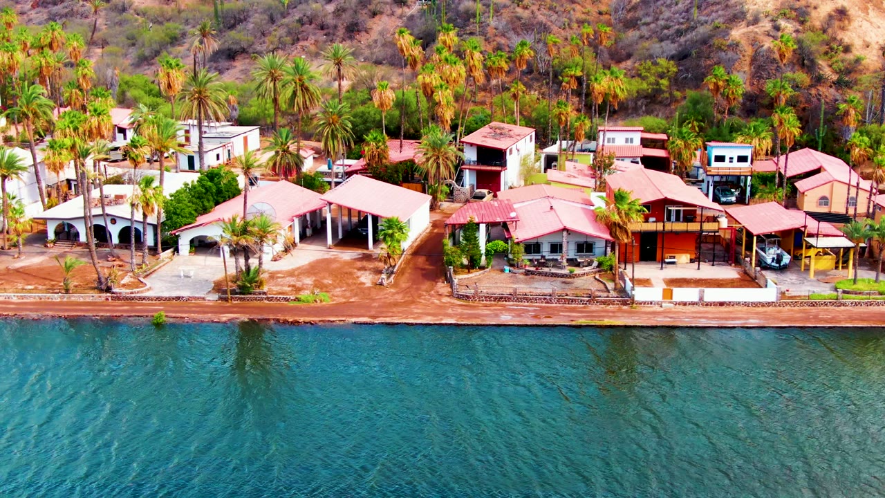 Mulege aerial frame with blue-green water across the lower half, red-roofed homes and a sandy bank through the center, and palms rising against a dry hillside with no visible people.
