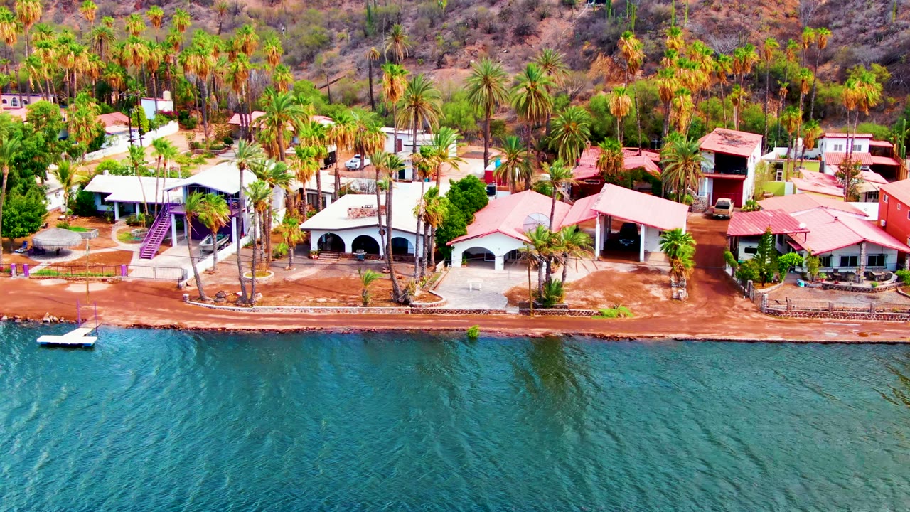 Opening frame of Mulege, Baja California Sur shoreline with blue-green water in front, low residential buildings and palms along the shore, dry hills behind, and no visible people.