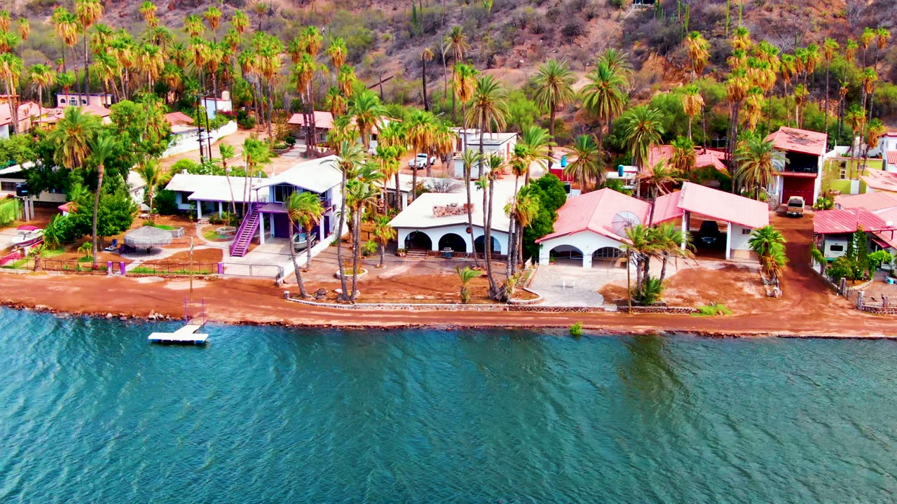 Mid-clip view of Mulege water and shore, with a small white boat near the lower left, pale and red-roofed buildings along the bank, dense palms, dry hill slope, and no visible people.