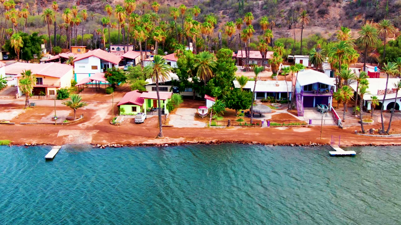 Aerial frame over calm blue water in Mulege, Baja California Sur, showing red-roof homes, palms, a sandy shoreline, a small dock, hillside behind, and no visible people.