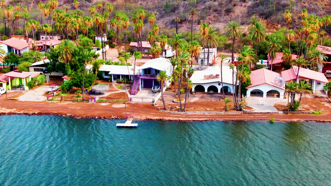 Early aerial frame over Mulege, Baja California Sur showing turquoise water, reddish shoreline, low buildings, tall palms, dry hillside, and no visible people.