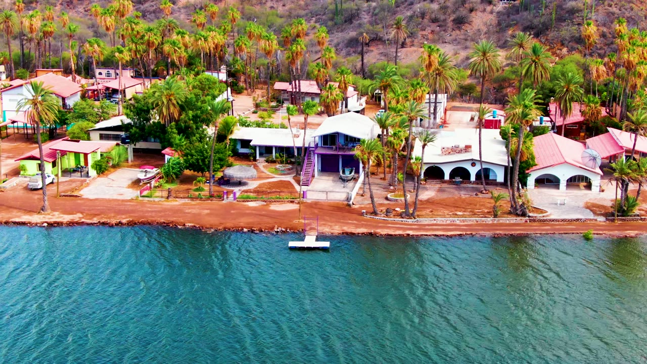 Mulege aerial frame showing red-roofed and white-roofed coastal structures behind a reddish shore, teal water in front, palms across the background, and no visible people.