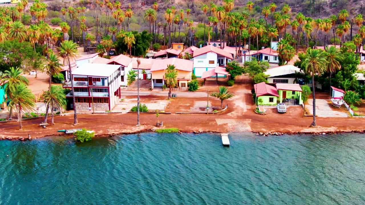 Aerial frame over Mulege, Baja California Sur, showing rippling blue water below a dirt shoreline, small waterfront houses, tall palms, dry hillside, and no visible people.