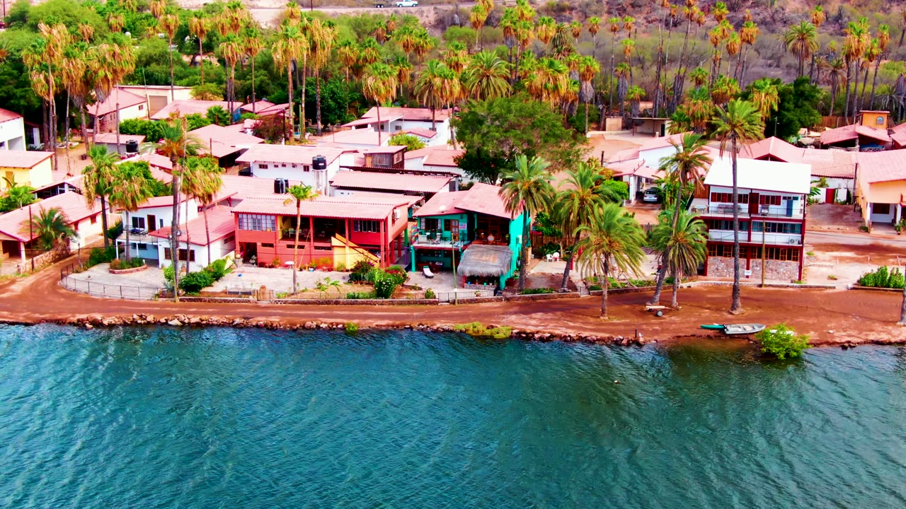 Aerial frame from Mulege, Baja California Sur showing rippled blue water below a reddish shoreline path, low waterfront homes, tall palms, dry brush beyond, and no visible people.