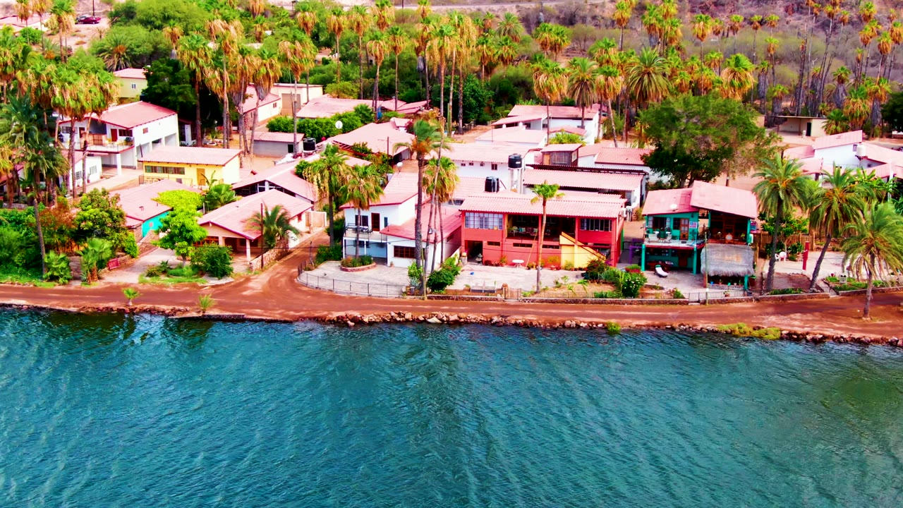 Aerial frame over Mulege, Baja California Sur showing blue-green water in front of a coastal road, low houses, and dense palms, with no visible people.