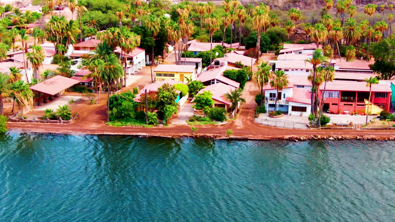 Opening aerial frame in Mulege, Baja California Sur showing rippling blue-green water, a narrow rocky edge, dirt road, palms, and colorful coastal homes with no visible people.