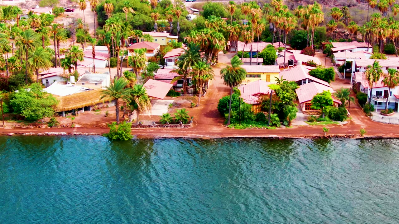 Mid-clip aerial frame over Mulege, Baja California Sur, showing textured blue water, a reddish dirt bank, clustered homes, tall palms, and no visible people.