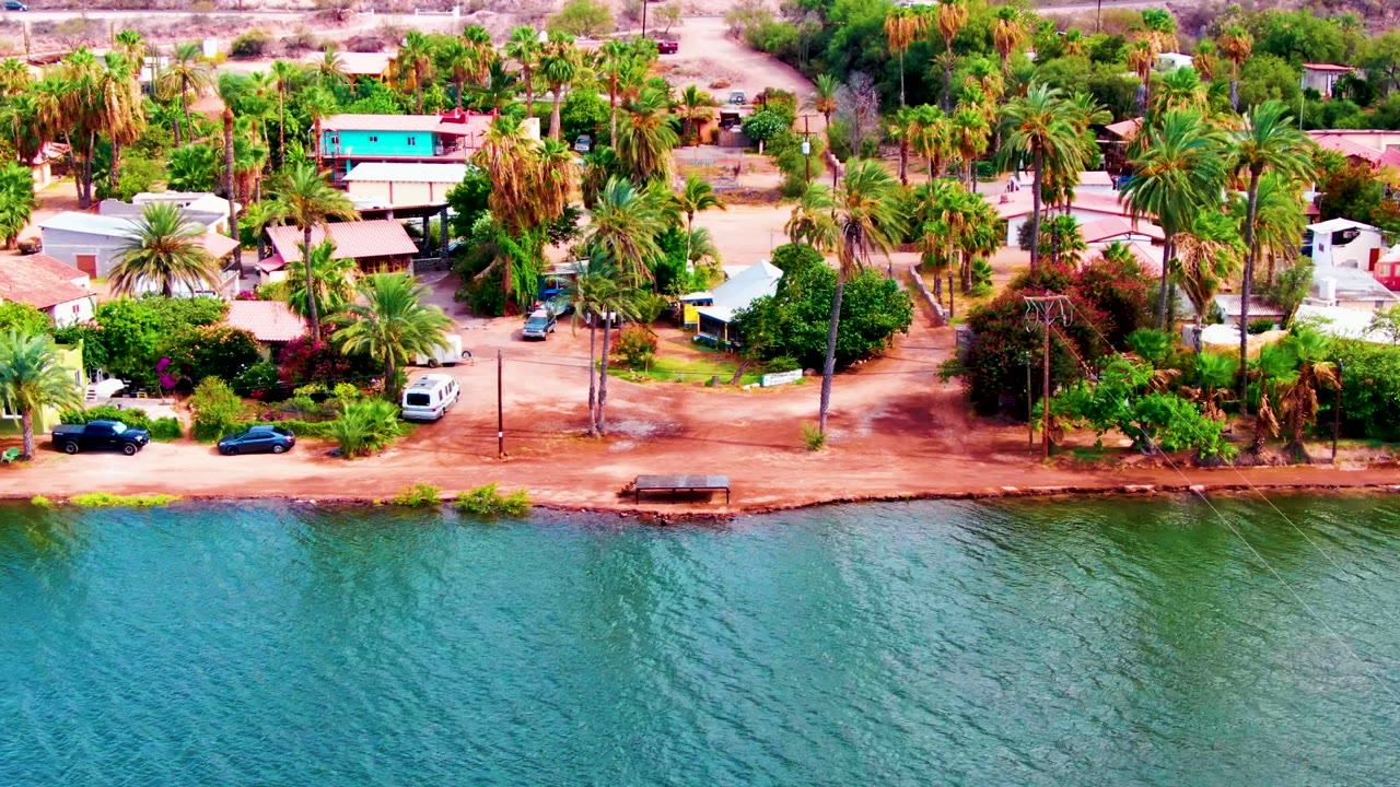 Aerial frame over Mulege, Baja California Sur, with rippling blue-green water below a reddish dirt shoreline, parked cars, homes, palms, and no visible people.