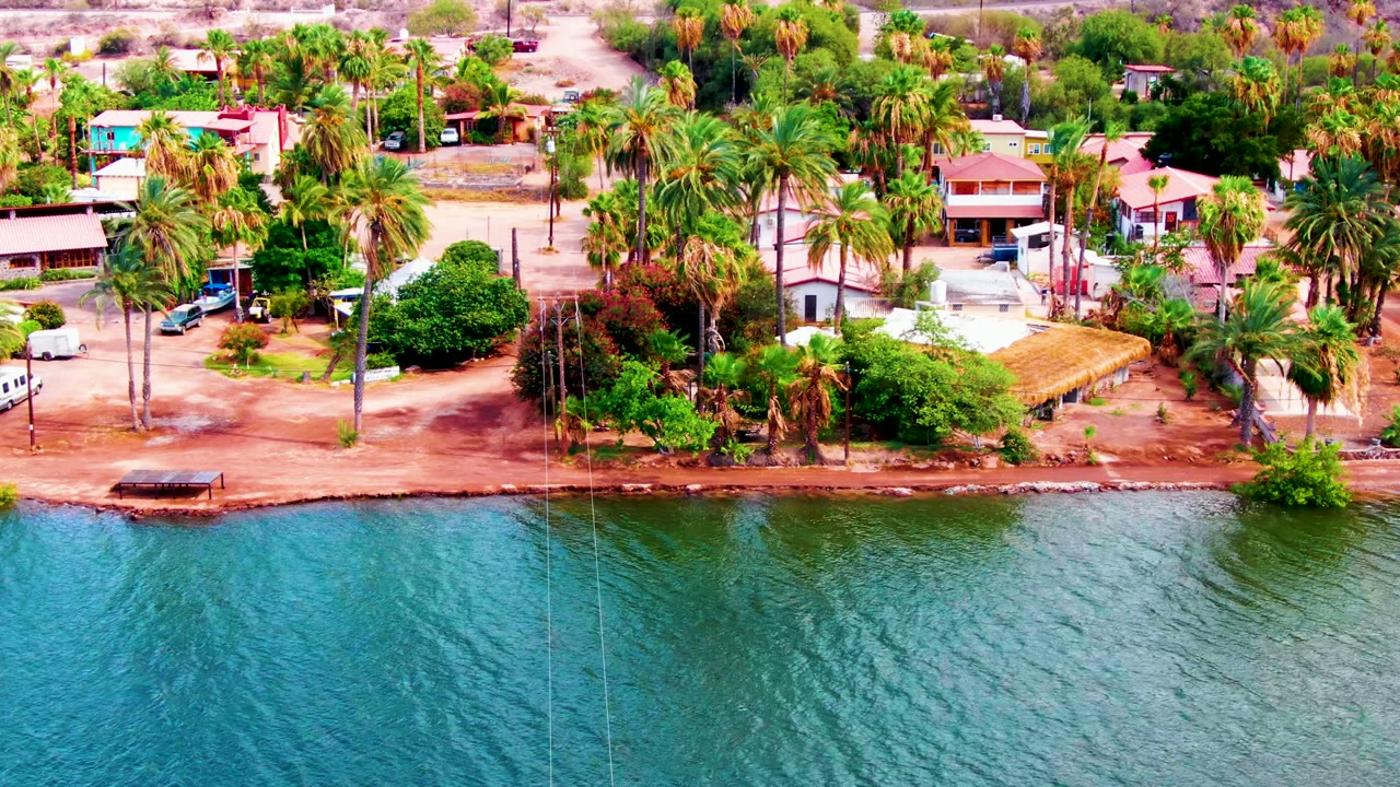 At 0:00, the Mulege aerial view frames blue-green water below a reddish waterfront road lined with palms, homes, parked vehicles, and no visible people.