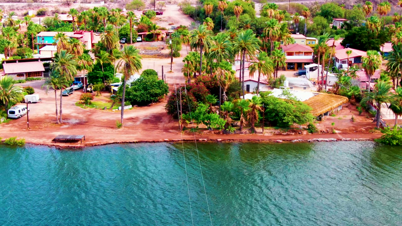 At 0:01, the drone-style view shows the Mulege waterfront road with a white van near the palms, a dark car on the road, houses behind, and no visible people.