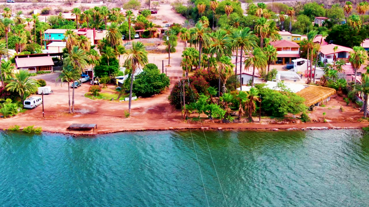 At 0:02, Mulege homes with pale and turquoise roofs sit behind tall palms while the road borders bright blue water, with no visible people.