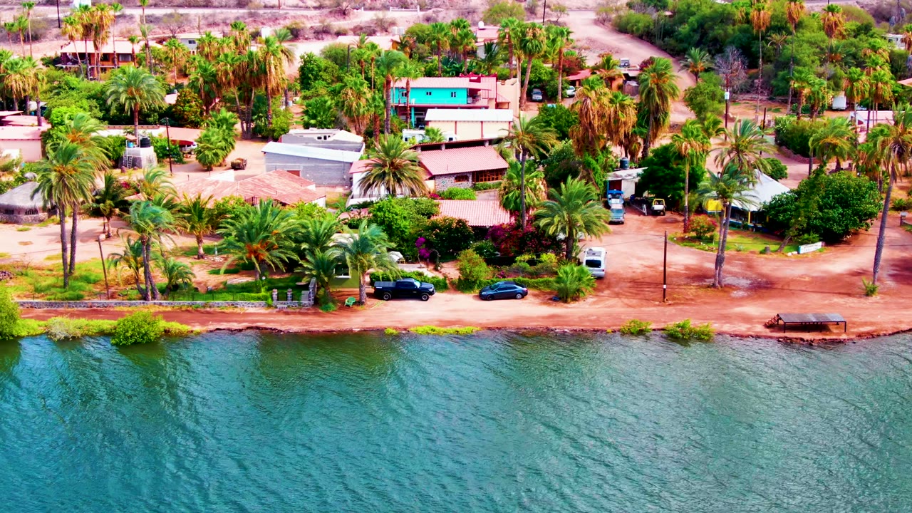 At 0:00, an elevated view in Mulege, Baja California Sur shows blue-green water below a dirt waterfront road lined with palms, small homes, parked vehicles, and no visible people.
