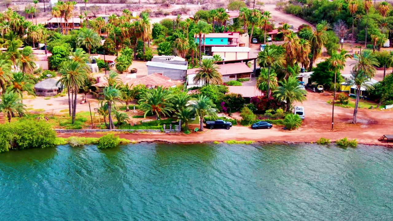 At 0:02, the aerial composition shows Mulege homes and palms above a dirt waterfront road, with textured blue-green water filling the lower half and no visible people.