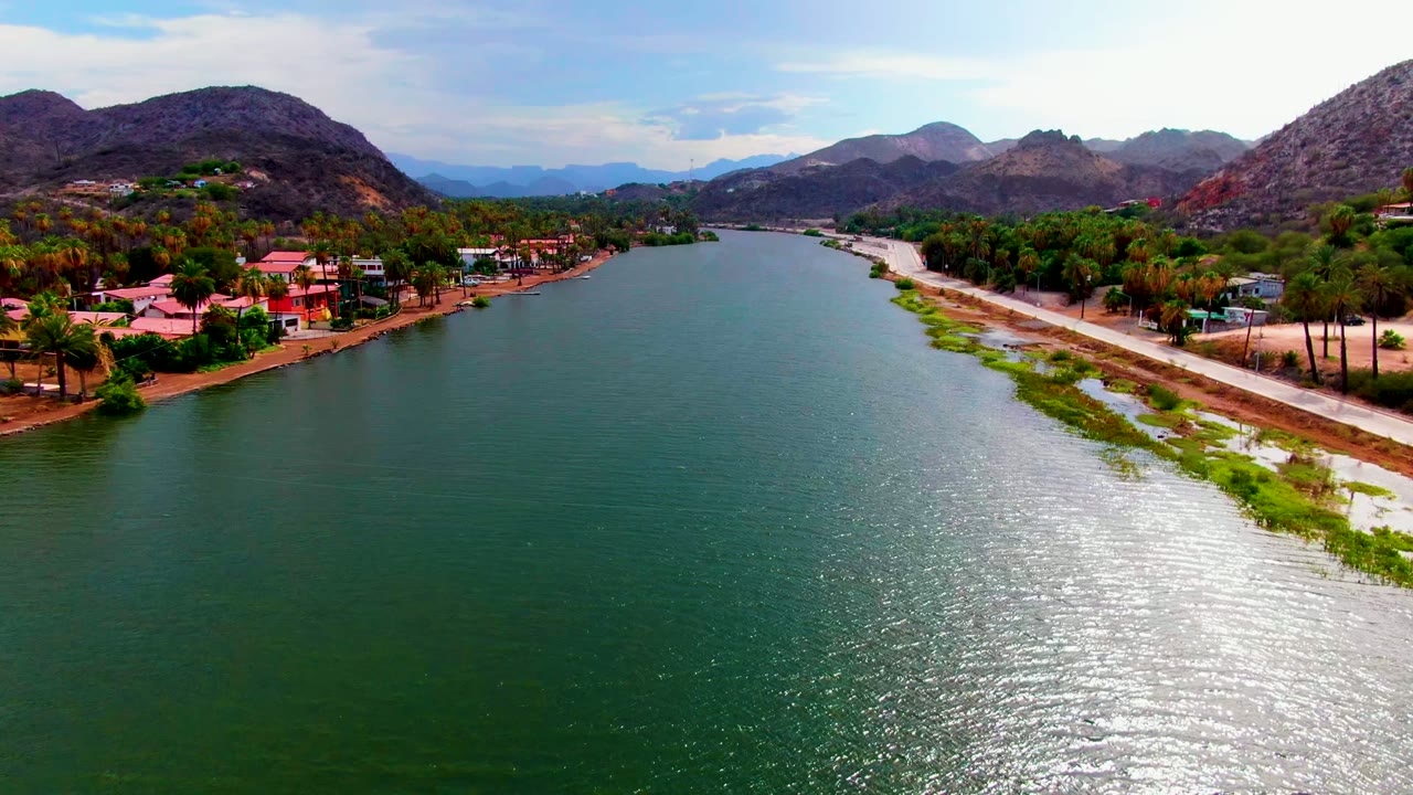 Opening aerial frame over the Mulege river, with green-blue water in front, homes and palms along the left bank, a pale road and shoreline on the right, mountains beyond, and no visible people.