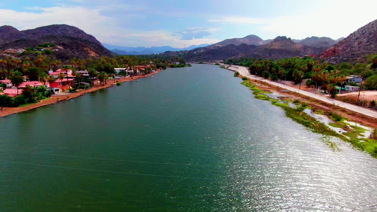 Mid-clip aerial frame over Mulege, showing a broad green river surface, bright ripples near the lower right, roadside vegetation on the right bank, red-roofed buildings on the left, hills behind, and no visible people.