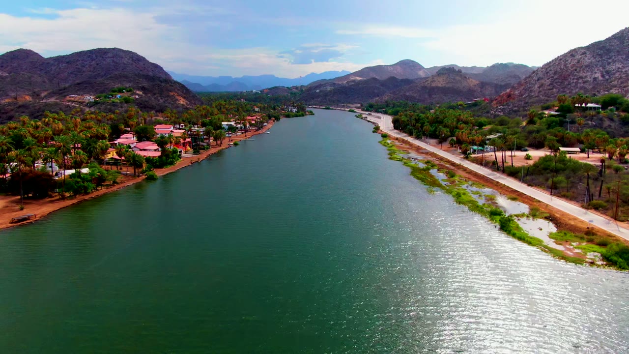 Aerial view in Mulege, Baja California Sur, showing a green-blue river beside a pale road, homes, palms, rocky mountains, and no visible people.