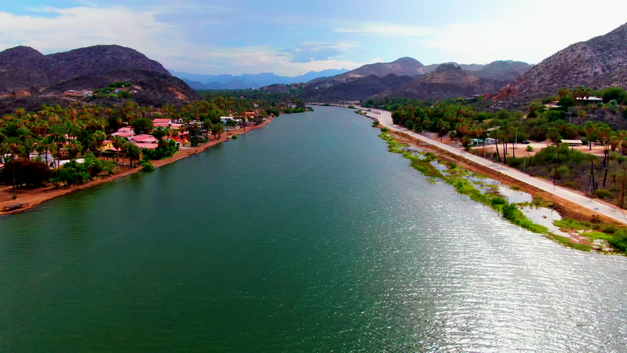 Wide aerial view over the Mulege river, with the road and green shoreline strip on the right bank, homes among palms, hills beyond, and no visible people.
