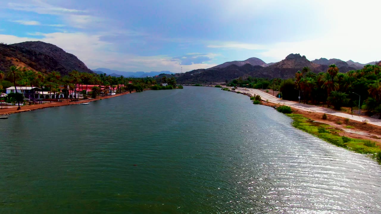 Opening frame of the Mulege, Baja California Sur river, with broad green-blue water in front, a pale road on the right shore, red-roofed buildings, palms, mountains, and no visible people.