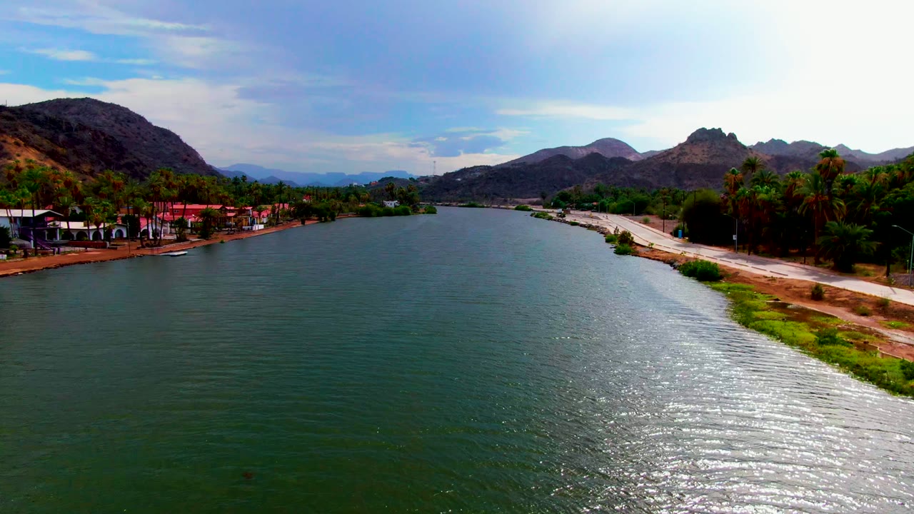 Aerial view down a river with bright water reflection and mountains beyond.