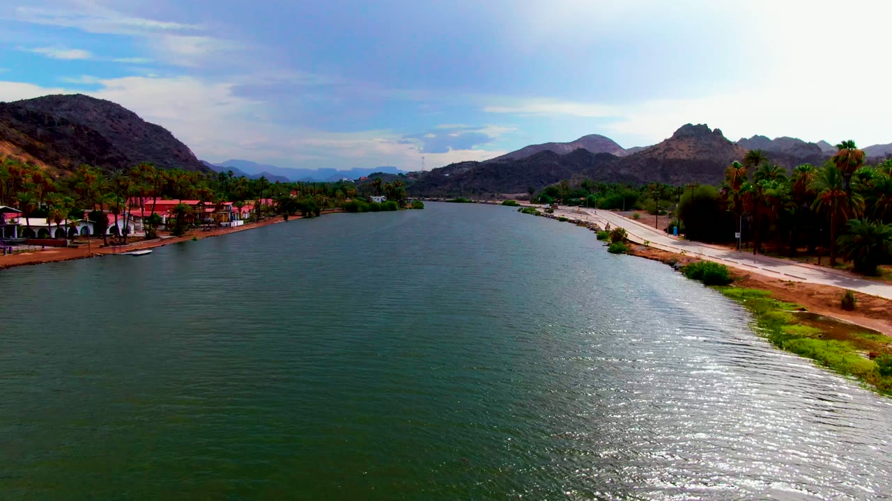 At 0:02, the Mulege river stretches toward dry mountains, with a clearer right-bank road, palms and buildings along the shore, bright sky reflection on the water, and no visible people.