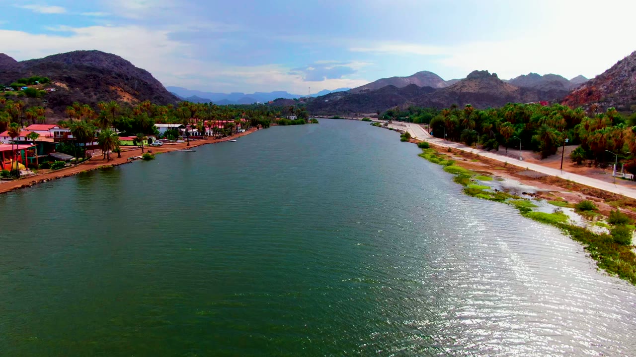 A wide aerial view in Mulege, Baja California Sur shows green-blue river water, a road and pale bank to the right, palm trees and small buildings to the left, rocky mountains beyond, and no visible people.