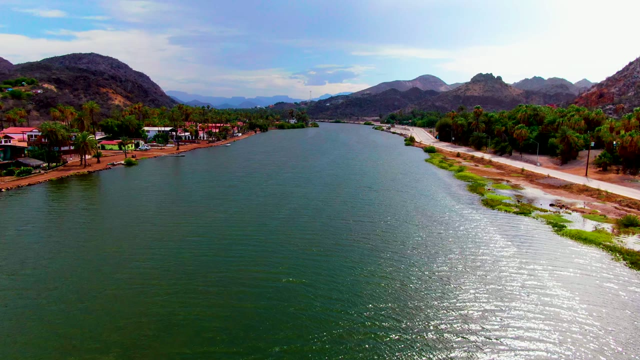 River channel between tree-lined shore and road with mountains beyond