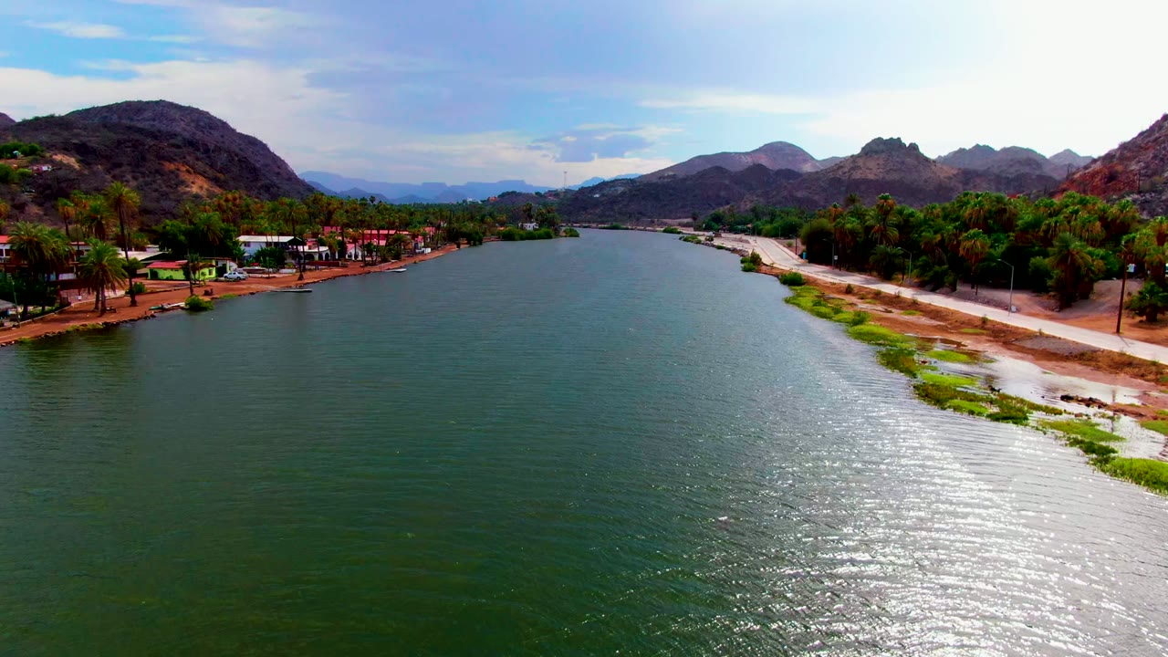 A wide aerial view in Mulege shows the river broadening in the foreground, bright sun glare across the water, a road and green shoreline plants to the right, palms and buildings to the left, and no visible people.