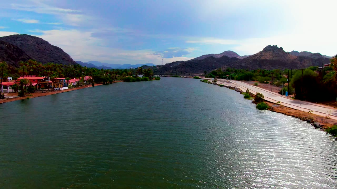 Aerial frame over the Mulege river corridor in Baja California Sur, with green-blue water in front, red-roofed buildings on the left bank, a road on the right bank, mountains beyond, and no visible people.