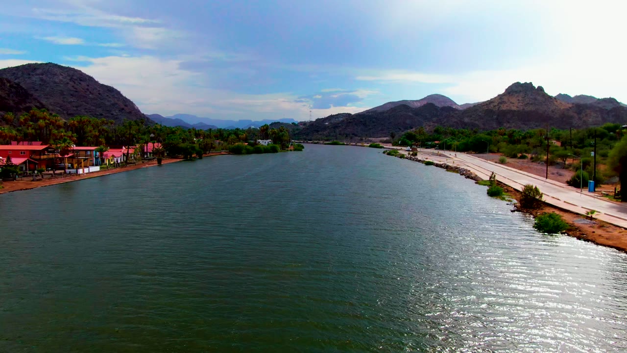 Drone-height frame over Mulege, Baja California Sur, with the river centered, red roofs and palms along the left edge, the road tracing the right shoreline, distant mountains, and no visible people.
