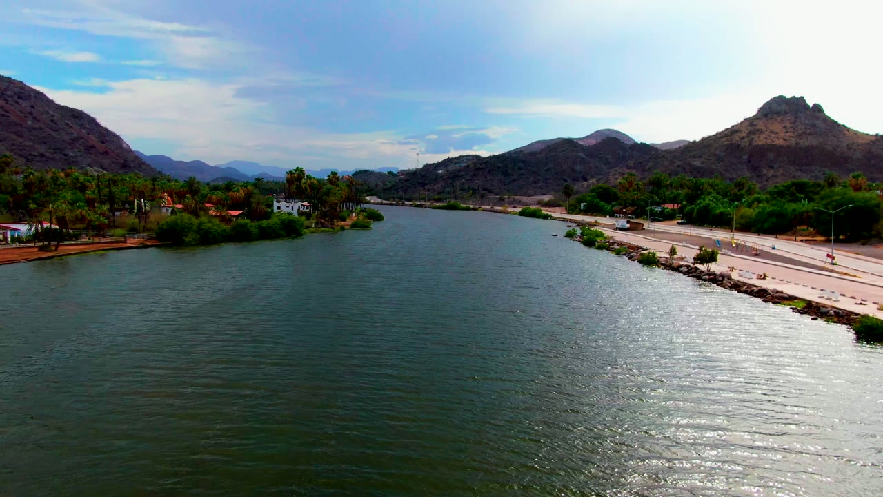 Aerial view in Mulege, Baja California Sur, with green river water in front, a road along the right bank, palms and buildings on the left bank, hills behind, and no visible people.