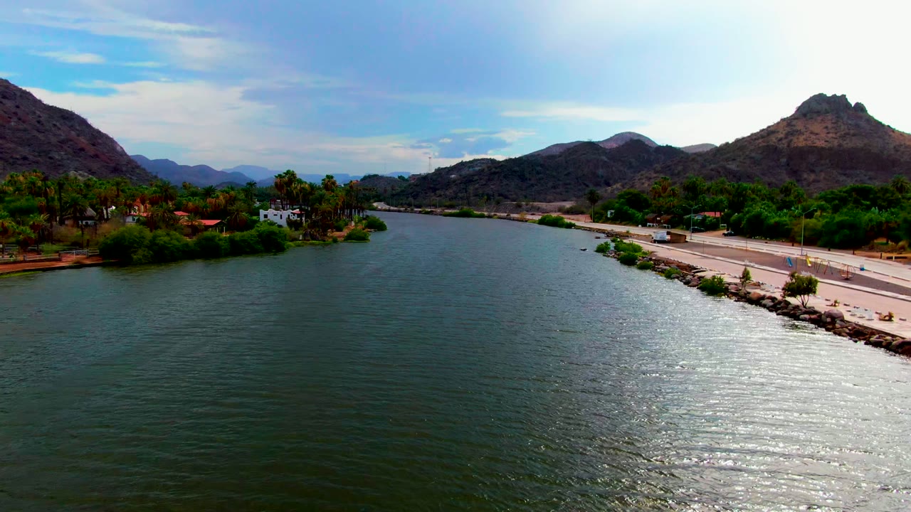 Aerial view in Mulege with the river widening across the foreground, the paved road edging the right side, palms and low buildings near the opposite bank, brown hills, and no visible people.