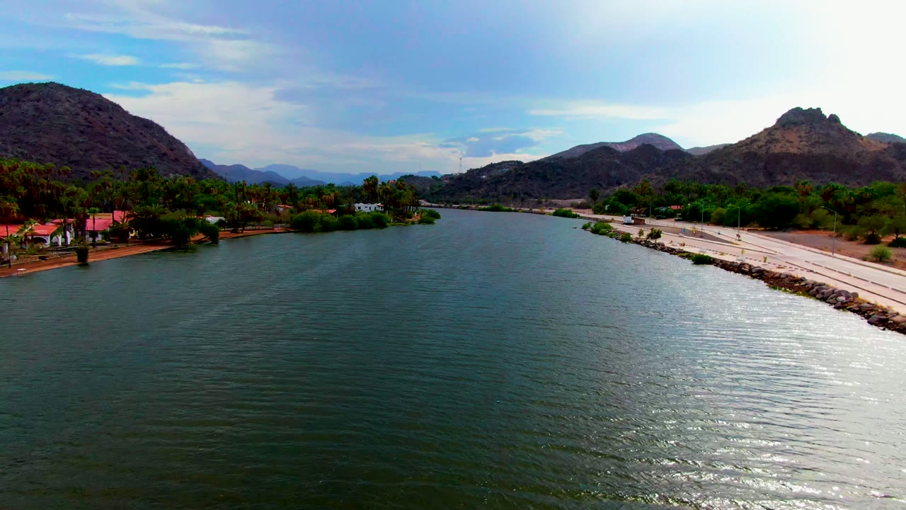 Opening aerial frame over Mulege, Baja California Sur, with green-blue river water in front, a road on the right bank, palms and low buildings along shore, mountains behind, and no visible people.