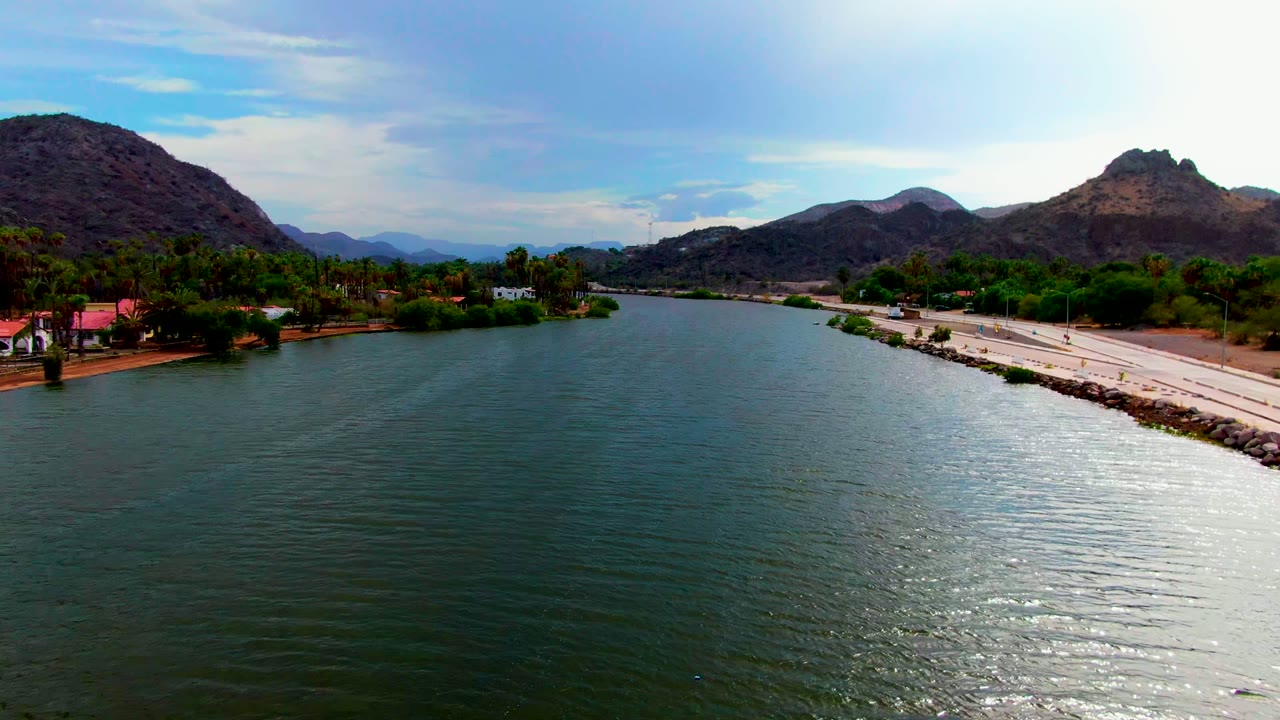 Wide aerial view of river water, roadside vehicles, palms, and mountains.