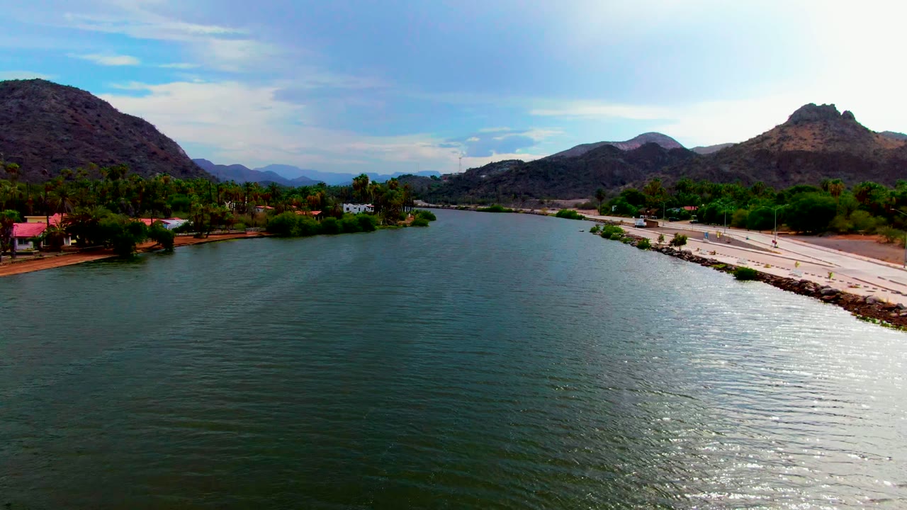 Aerial view over Mulege riverbank with the road and rocky shoreline on the right, palms and small structures near the water, layered mountains in the distance, and no visible people.