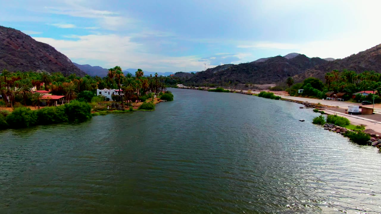 Opening aerial frame over a wide river in Mulege, Baja California Sur, with palm trees, small buildings, a roadside bank, brown mountains, bright sky, and no visible people.