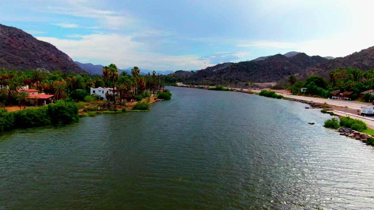 River aerial with shoreline palms, white building, road, and mountain ridge.