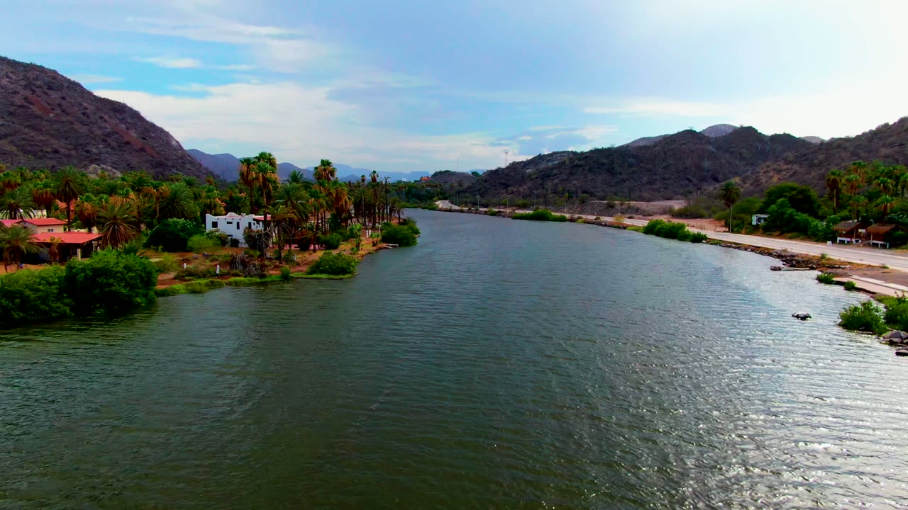 Third frame looks along the Mulege river channel with bright water reflection on the right, palm trees and low buildings near the bank, a road tracing the shoreline, hills, and no visible people.